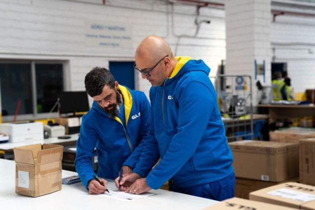 Two GLS delivery workers review and sign shipment documents in a logistics warehouse surrounded by parcels.