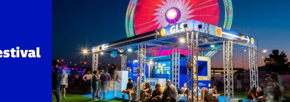 Stand nocturno de GLS iluminado en Mad Cool Festival, con asistentes descansando y noria iluminada de fondo