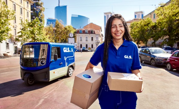 Hymyilevä GLS-työntekijä kävelee kameraa kohti kaksi pakettia käsissään ja taustalla on sähköinen GLS-jakeluauto