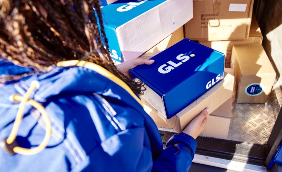 GLS-kuljettaja kuvattuna takaa ja hän pitää käsissään GLS-paketteja, jotka ovat GLS-pakettiauton kyydissä