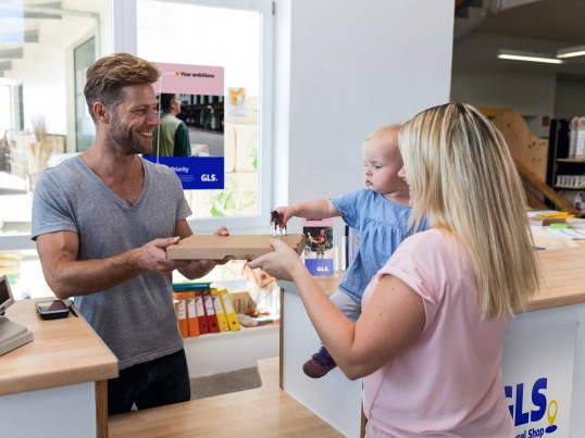 Une femme avec un bébé récupère un colis auprès d’un commerçant dans un Point Relais GLS.