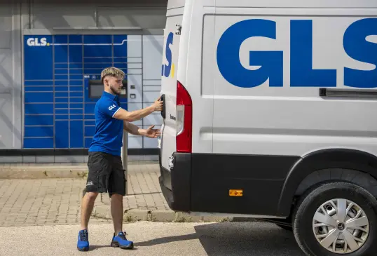 Livreur GLS en polo bleu ouvre la porte arrière d'un utilitaire GLS blanc, devant des consignes à colis GLS.