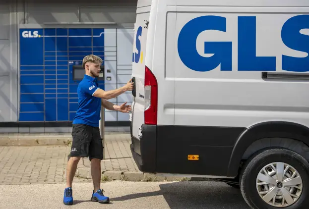 Livreur GLS en polo bleu ouvre la porte arrière d'un utilitaire GLS blanc, devant des consignes à colis GLS.