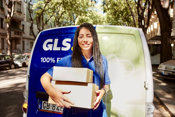 A smiling GLS delivery driver holds two parcels in front of a parked GLS delivery van on a tree-lined city street. Residential buildings and parked cars are visible in the background.