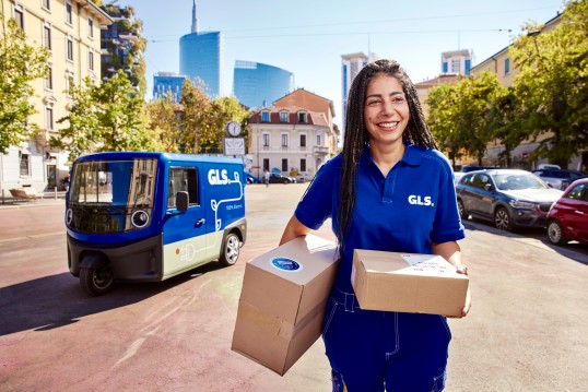 GLS delivery driver smiling while carrying parcels in front of a GLS delivery vehicle on a city street.