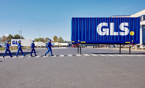 GLS team walking across a secured pedestrian crossing in front of a blue semi-trailer at a parcel logistics distribution center.