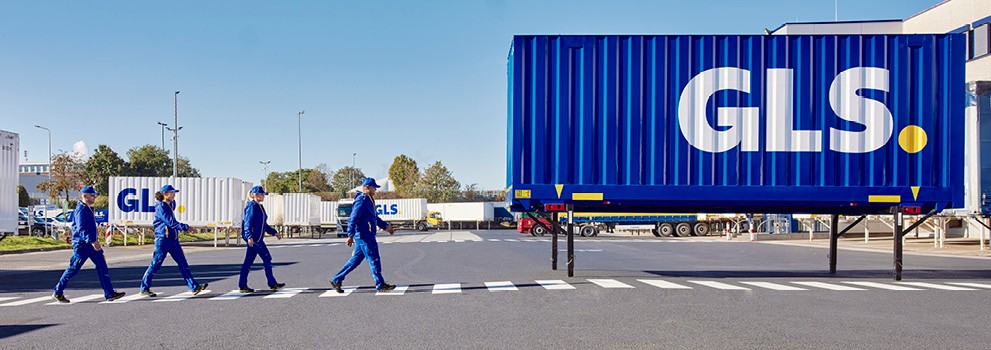 GLS team walking across a secured pedestrian crossing in front of a blue semi-trailer at a parcel logistics distribution center.