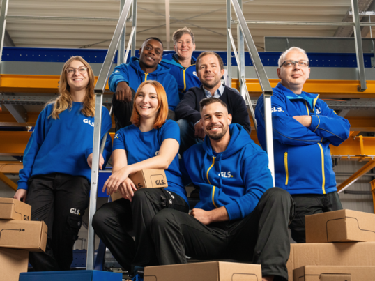 Team of GLS employees in professional uniforms inside a logistics warehouse, surrounded by parcels, illustrating career opportunities and logistics jobs.