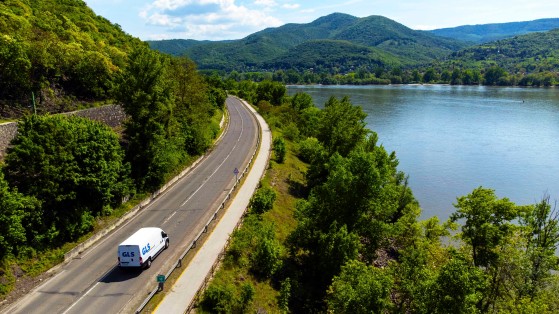 A GLS delivery van drives along a winding road beside a wide river, surrounded by lush greenery and forested hills. The scenic landscape stretches beneath a partly cloudy sky.