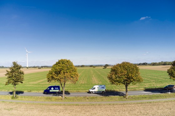 Two GLS delivery vans drive along a tree-lined country road through expansive farmland. A wind turbine stands in the distance beneath a clear blue sky, while a car travels farther along the same road.