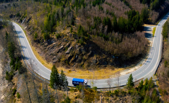 Blue GLS semi-trailer truck on a winding mountain road through a forested landscape.