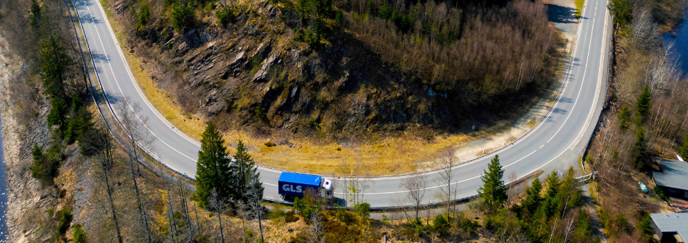 Blue GLS semi-trailer truck on a winding mountain road through a forested landscape.