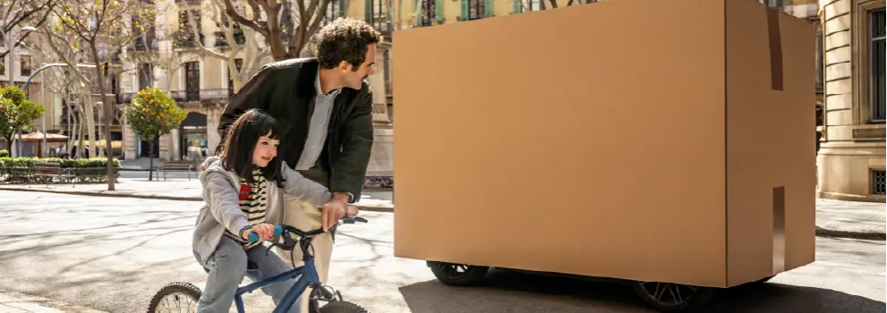 Ein Vater betrachtet ein großes Paket, während er seiner Tochter das Fahrradfahren beibringt.