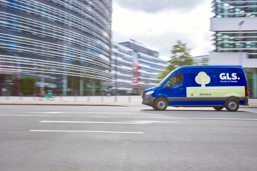 A blue GLS delivery van drives along a city street in front of modern glass office buildings.