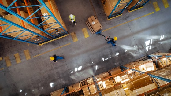 Overhead view of two warehouse workers performing separate tasks among aisles of stacked boxes.